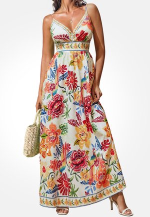 Femme portant une robe maxi blanche sans manches avec des motifs floraux et de feuilles colorés, tenant un sac à main tissé, et portant des talons blancs.