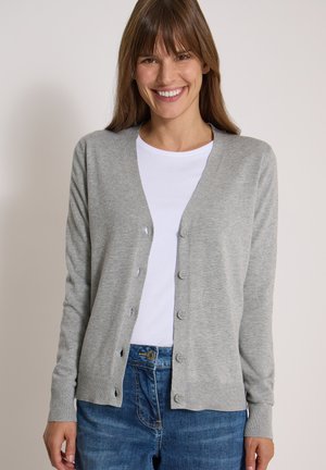 Femme souriante portant un cardigan gris boutonné sur une chemise blanche et un jean bleu sur un fond clair uni.