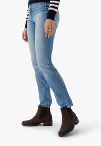 Person som bär ljusblå jeans, mörkbruna mockaskor med ankellängd och en marinblå randig långärmad topp, stående på en vit bakgrund.