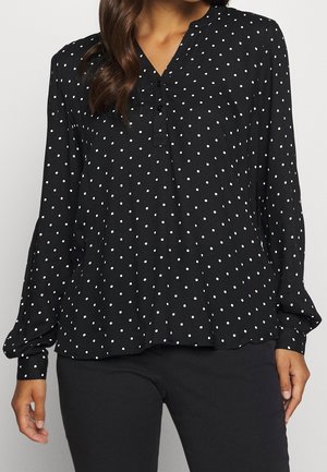 Blouse noire à manches longues avec des pois blancs, boutonnée au niveau de l'encolure, portée avec un pantalon noir.