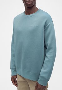 Homme portant un sweat-shirt bleu clair à col rond avec poignets et ourlet côtelés, associé à un pantalon beige, debout devant un fond blanc uni.
