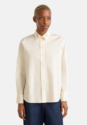 Camicia button-up color crema con un design ricamato a foglia sul colletto. Maniche lunghe, texture liscia e vestibilità rilassata. Indossata con jeans blu scuro.