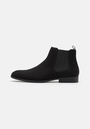Bottine Chelsea noire en daim avec panneaux latéraux élastiques, petit talon empilé, bout arrondi et languette à l’arrière.
