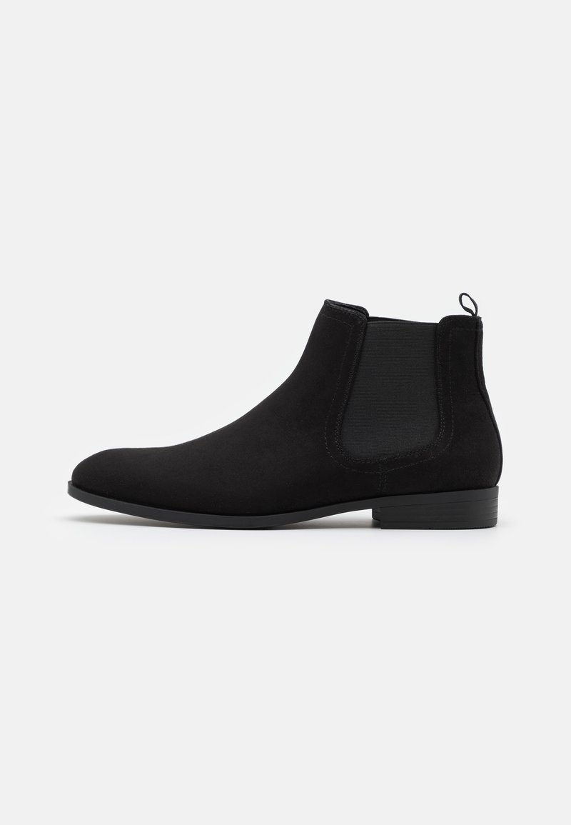 Bottine Chelsea noire en daim avec panneaux latéraux élastiques, petit talon empilé, bout arrondi et languette à l’arrière.