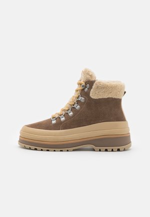 Braune Wildleder-Wanderstiefel mit cremefarbenem Fellrand, Metallösen und hellbrauner Gummisohle. Robustes, klobiges Design mit Schnürung vorne.