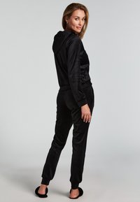 Musta samettinen urheilupuku, jossa on hupparitoppi ja suppilomaiset housut. Pehmeä rakenne ja joustavat hihansuut. Malli seisoo selkä kameraan päin.