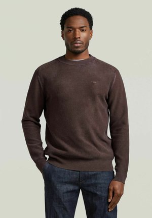 Uomo con maglione a coste marrone scuro e jeans blu, in piedi con una mano in tasca contro uno sfondo chiaro e uniforme.