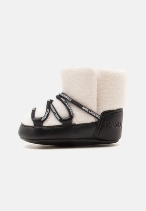 Pequeña bota blanca de forro polar con suela negra y correas negras con la marca "Karl Lagerfeld", mostrada de lado sobre un fondo blanco.