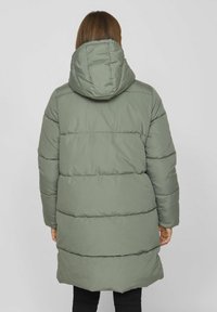 Doudoune verte avec capuche, présentant des sections matelassées et une coupe longue. Le tissu a une texture lisse et elle est portée sur un pantalon noir.