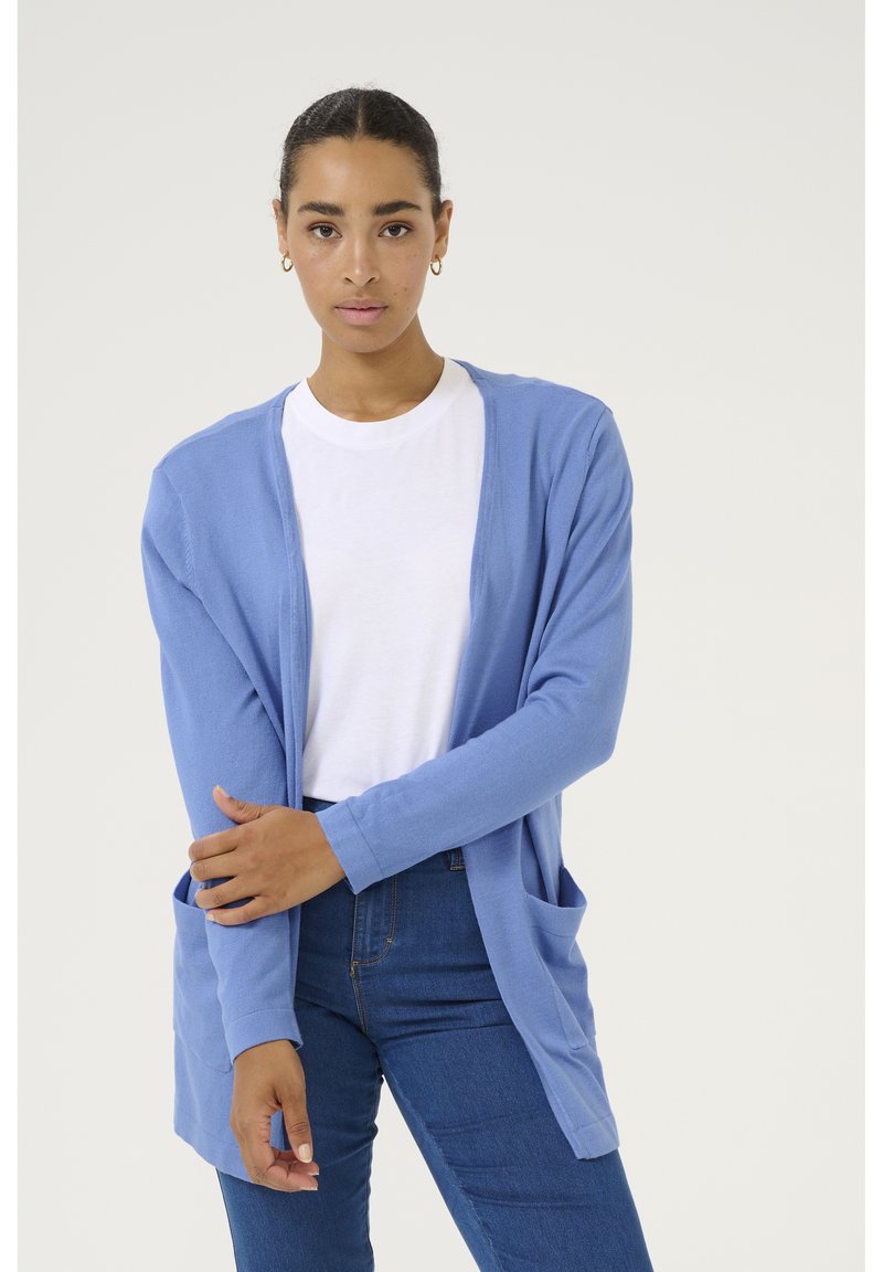 Cardigan bleu clair à manches longues et à l'avant ouvert. Comprend deux poches avant et une coupe décontractée. Porté par-dessus un t-shirt blanc et un jean bleu.