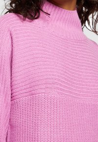 Rosa gestrickter Pullover mit hohem, geripptem Kragen und durchgehendem Muster aus horizontalen und vertikalen Strukturen. Weiches und gemütliches Aussehen.
