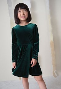 Ung pige med glat sort hår iført en langærmet mørkegrøn fløjls kjole, stående mod en lys baggrund med cirkulære hængende dekorationer.