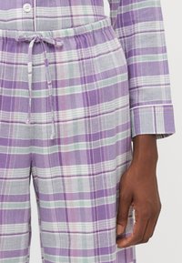 Fialové, biele a zelené kockované nohavice s elastickým pásom, nosené s rovnakým dlhým rukávom, vedľa ktorých odpočíva tmavšia pleť rúk.