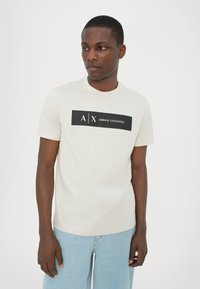 Lys beige bomull t-skjorte med en svart rektangulær logo som har «A|X ARMANI EXCHANGE» tekst sentrert på brystet. Rund hals, korte ermer.
