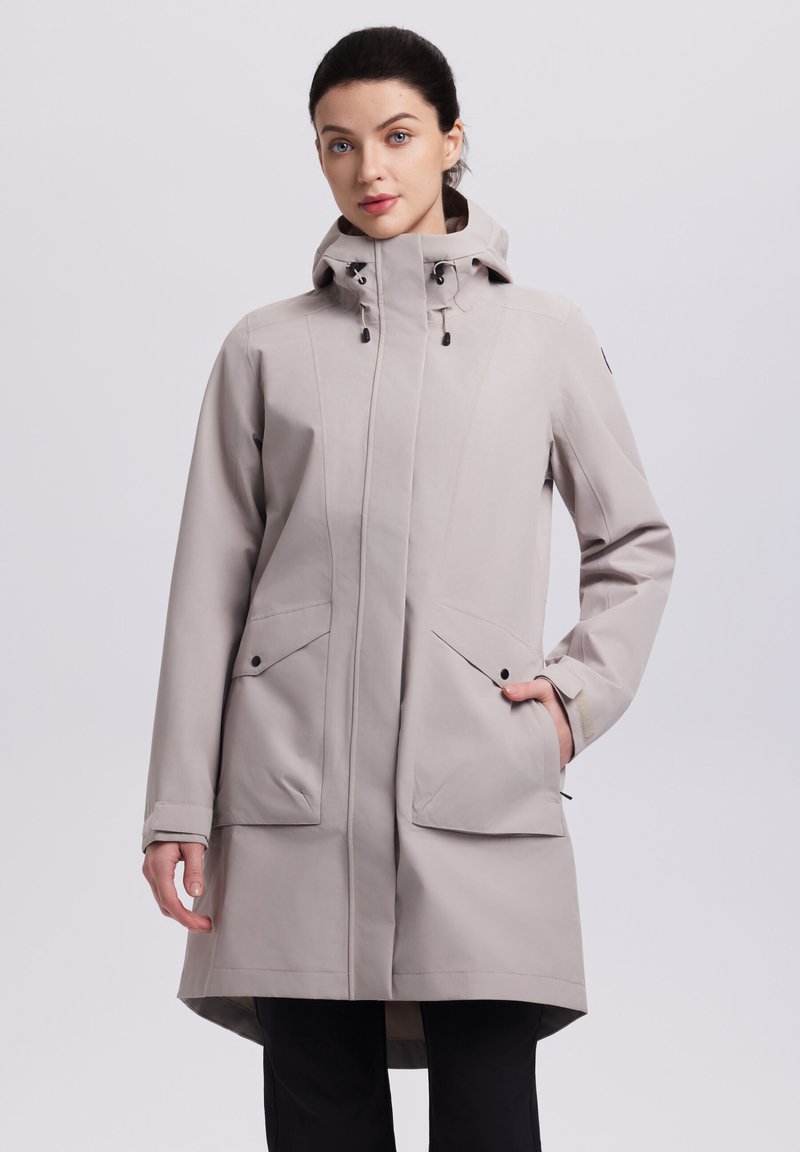 Mujer con gabardina impermeable beige claro con capucha y bolsillos delanteros con solapa, y pantalones negros, de pie frente a un fondo liso.