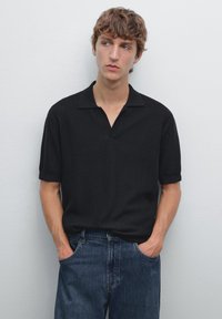 Giovane uomo con capelli scompigliati, che indossa una polo nera e jeans, in piedi contro un muro bianco uniforme, con le mani in tasca, che guarda leggermente di lato.