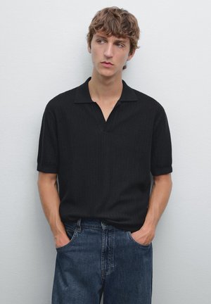 Jonge man met warrig haar, gekleed in een zwart polo shirt en jeans, staand tegen een witte muur, met zijn handen in zijn zakken, lichtjes wegkijkend.