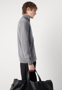 Junger Mann in grauem Zip-Up-Pullover und schwarzer Hose hält eine große schwarze Ledertasche vor einem einfarbigen Hintergrund.