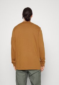 Osoba s tmavými vlasy v drdolu, oblečená v hnědé dlouhé košili a olivově zelených kalhotách, stojící proti obyčejné bílé zdi.