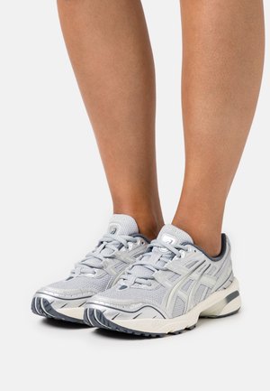 Baskets de sport gris clair avec une tige en maille texturée, des accents réfléchissants et des semelles en caoutchouc noir, présentant un design à lacets et un talon rembourré.