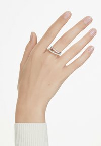 Silberring mit verschlungenem Design, versehen mit minimalistischen Linien und kleinen klaren Akzenten. Auf einer Hand vor einem schlichten Hintergrund platziert.