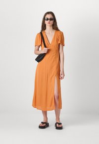 Orangenes Midi-Kleid mit kurzen Puffärmeln, tiefem V-Ausschnitt und seitlichem Schlitz. Kombiniert mit schwarzen Sandalen und einer kleinen Umhängetasche.
