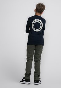 Maglietta a maniche lunghe blu marina con grafica circolare bianca sulla schiena, abbinata a pantaloni cargo verde oliva e sneakers nere e bianche.