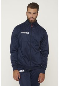 Chaqueta deportiva azul marino de tejido suave con cuello alto, cierre completo y logo blanco "LEGEA" en el lado izquierdo del pecho. Tirador de cremallera rojo.