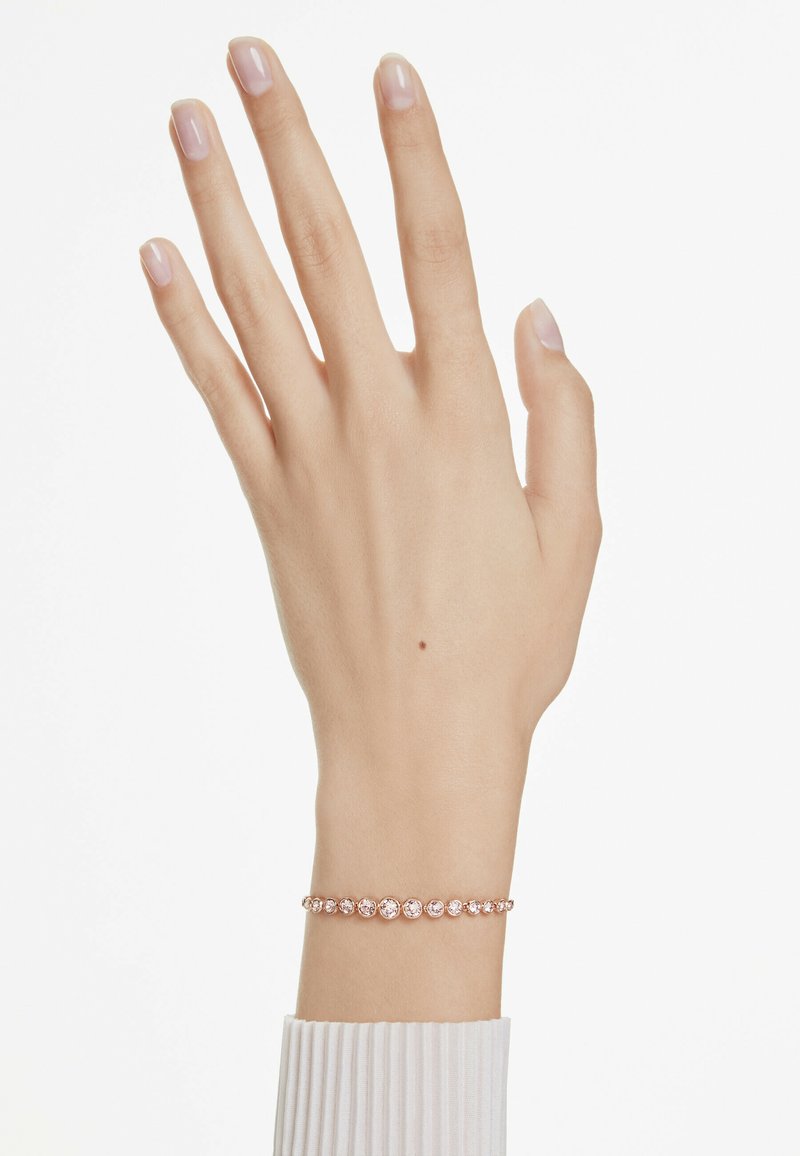 Pulsera de oro rosa con múltiples piedras claras dispuestas en línea. Mano con uñas ligeramente cuidadas mostradas sobre un fondo blanco.