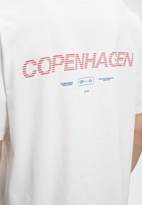 Άσπρη μπλούζα με κόκκινα ριγέ γράμματα "COPENHAGEN" και μικρό μπλε κείμενο και λογότυπα από κάτω στην πλάτη ενός ατόμου.