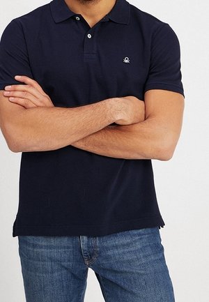 Man stands with arms crossed wearing a navy blue polo shirt and blue jeans against a plain background.