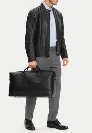 Mann in schwarzer Lederjacke, hellblauem Hemd und grauer Hose, der eine große schwarze Gino Rossi Reisetasche an den Griffen hält.