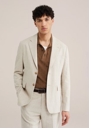 Jonge man die een crèmekleurig pak draagt met een bruin textuur polohemd en gevlochten riem, staand met één hand in de zak tegen een effen achtergrond.