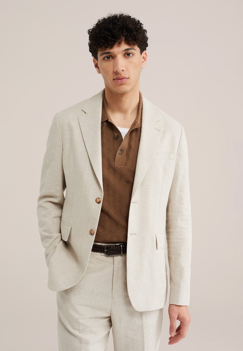 Jeune homme portant un costume crème avec un polo marron texturé et une ceinture tressée, debout avec une main dans la poche sur un fond uni.