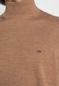 Brun polokrage med ribbad textur och figurnära design, med en liten svart "CK"-logotyp på vänster sida.