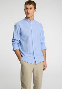Camisa de botón de manga larga azul claro con rayas verticales blancas, con cuello mandarín y mangas arremangadas. Combinada con pantalones beige.