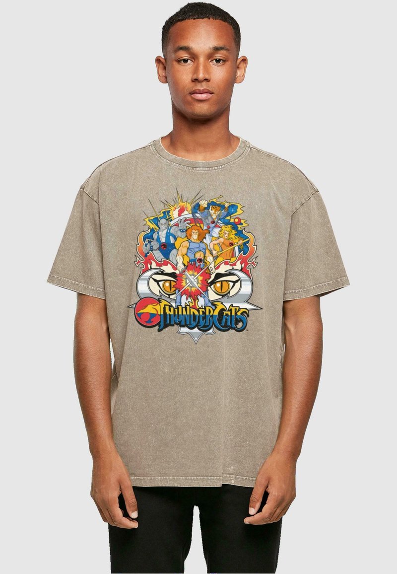 Tan spalvos per didelis marškinėliai su spalvingu "ThunderCats" grafikos spaudiniu priekyje, apvaliu kaklu ir trumpomis rankovėmis.