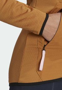 Main d'une personne insérée dans la poche zippée d'une veste marron avec bordure noire et tirette blanche, vue de côté.