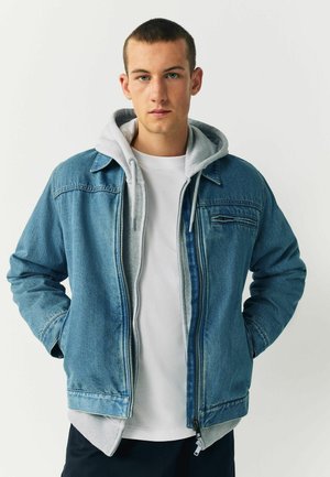 Jachetă din denim de culoare albastru deschis, cu fermoar la față, căptușeală gri de tip hoodie și buzunare laterale. Textură netedă cu un design clasic.