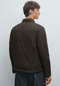 Young man facing sideways wearing a dark brown padded jacket and dark pants against a plain light gray background.
