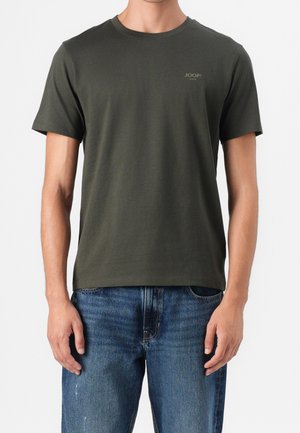 Homme portant un t-shirt uni vert foncé à manches courtes avec un petit logo "JOOP! JEANS" sur la poitrine, associé à un jean bleu en denim.