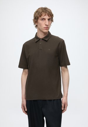 Ung mand med lyst krøllet hår iført en mørkebrun kortærmet poloshirt og sorte bukser mod en ensfarvet hvid baggrund.