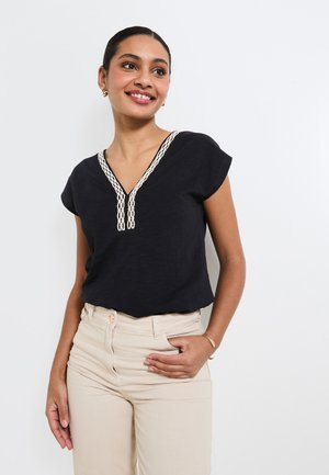 Haut noir à manches courtes avec un col en V, orné d'une bordure crème et d'une coupe décontractée. Porté avec un pantalon clair.