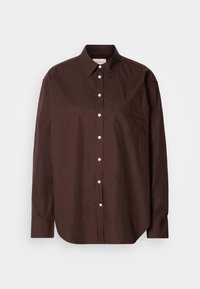 Chemise marron foncé à manches longues avec boutons blancs et poche poitrine gauche sur un fond clair uni.
