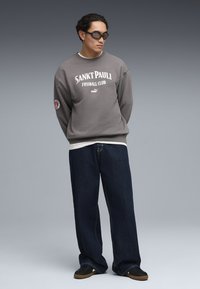 Sudadera gris con estampado "SANKT PAULI FUSSBALL CLUB", combinada con vaqueros sueltos azul oscuro y zapatillas negras. Cuello redondo, tela suave y mangas largas.