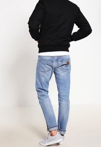 Mand iført lysblå opsmøgede jeans, hvide sneakers, hvid skjorte og sort jakke, stående med hænderne på hoften, vendt væk.
