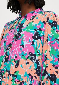 Donna che indossa una blusa con stampa astratta multicolore, con forme rosa, verde, teal, pesca e nera, dotata di colletto e bottoni lungo il davanti.