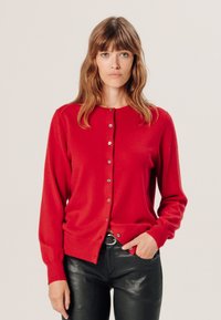 Roter Cardigan mit rundem Ausschnitt, langen Ärmeln und Knopfverschluss. Weicher Stoff, taillierte Form, kombiniert mit schwarzen, schmal geschnittenen Hosen.