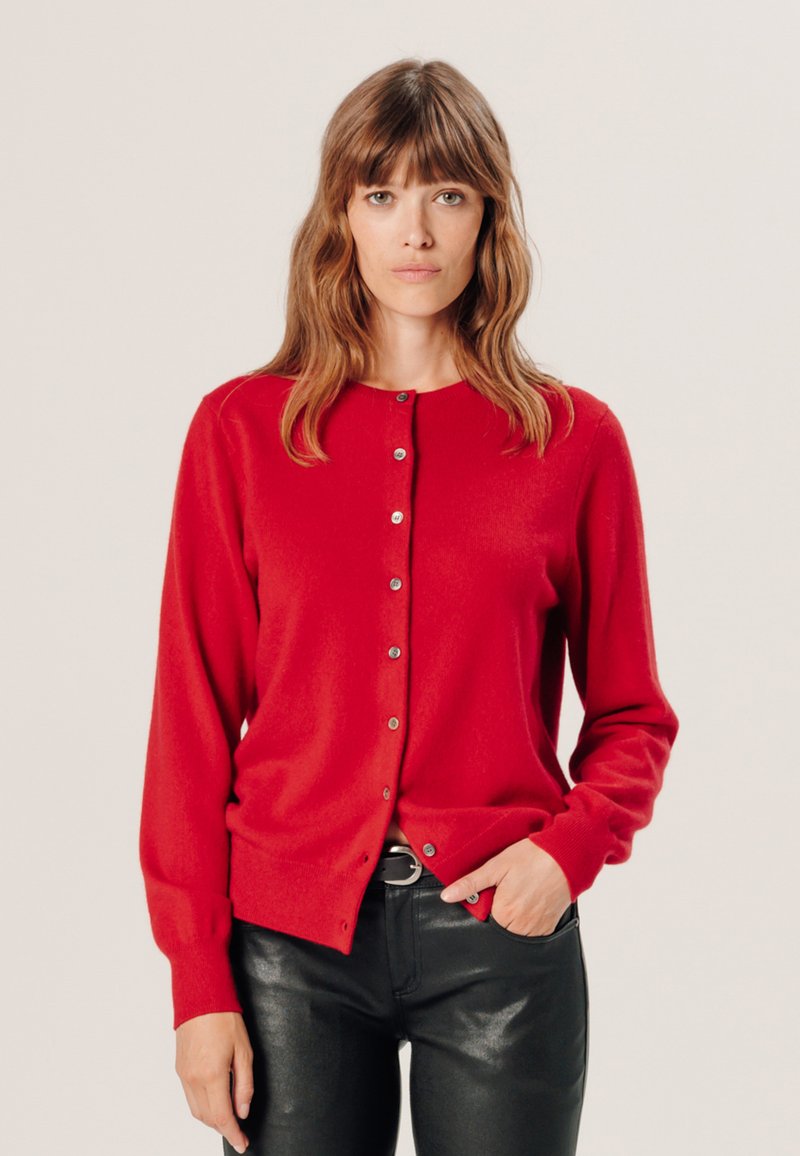 Roter Cardigan mit rundem Ausschnitt, langen Ärmeln und Knopfverschluss. Weicher Stoff, taillierte Form, kombiniert mit schwarzen, schmal geschnittenen Hosen.