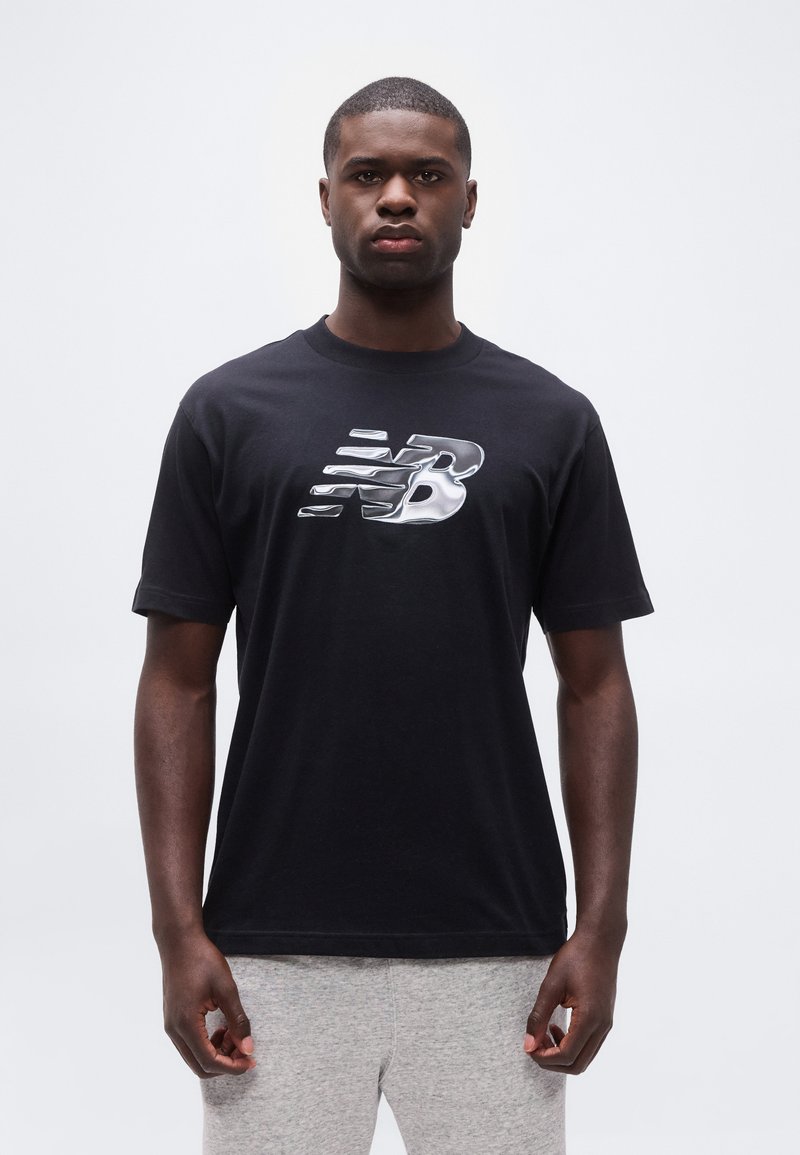 Νέος άνδρας που φοράει μαύρο T-shirt με ασημένιο στυλιζαρισμένο λογότυπο "NB" και ανοιχτό γκρι φόρμα, στέκεται μπροστά σε λευκό απλό φόντο.